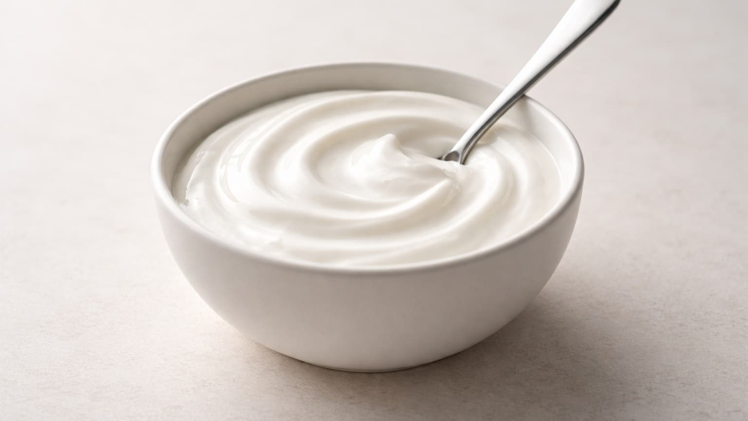 Minimalist food photograph shot from a 45-degree angle. A small matte white ceramic bowl on a light neutral stone surface contains thick white Greek yogurt mixed to a smooth, glossy consistency. The yogurt surface is uniform, creamy, and free of lumps or streaks. A plain metal spoon rests partially inside the bowl, indicating gentle stirring. Soft diffused daylight from the left, neutral tones, shallow depth of field, no props, no background elements.