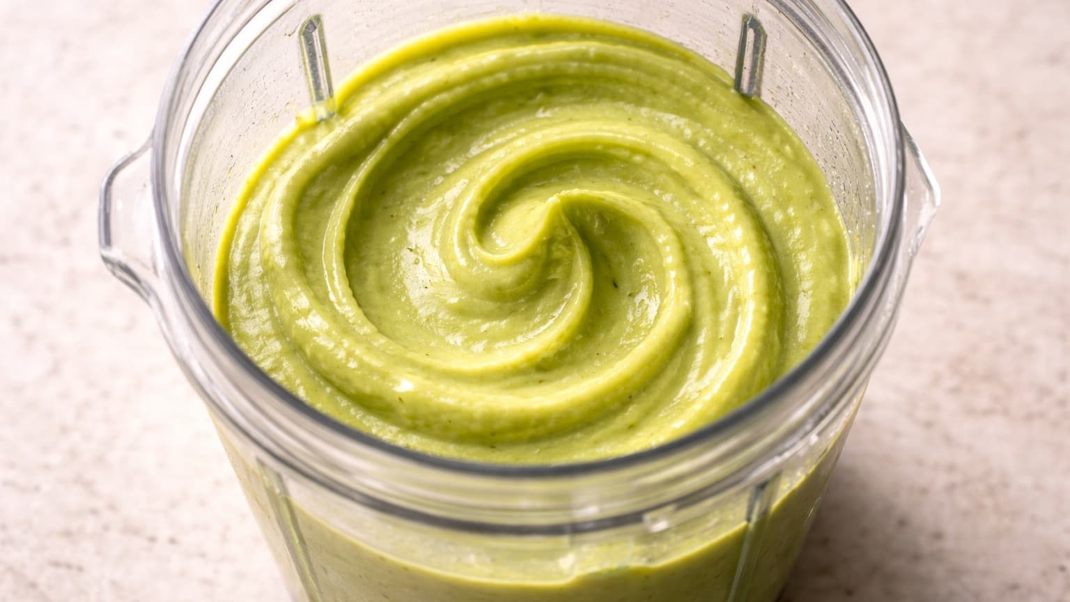Close-up food photograph shot from above at a slight angle. A high-speed blender cup filled with thick, smooth, vivid green avocado cream, surface glossy and uniform with no lumps. Soft daylight, neutral background, clean and realistic.