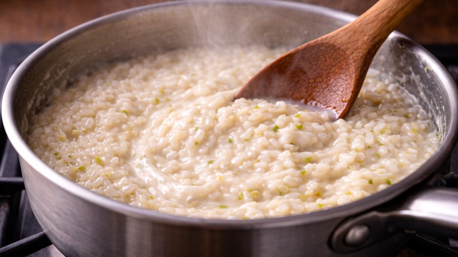 A pot of creamy risotto being stirred as white coconut milk blends into the rice. The mixture appears smooth, glossy, and pale ivory with tiny green flecks of lime zest. A wooden spoon creates soft swirls in the creamy texture while steam rises.