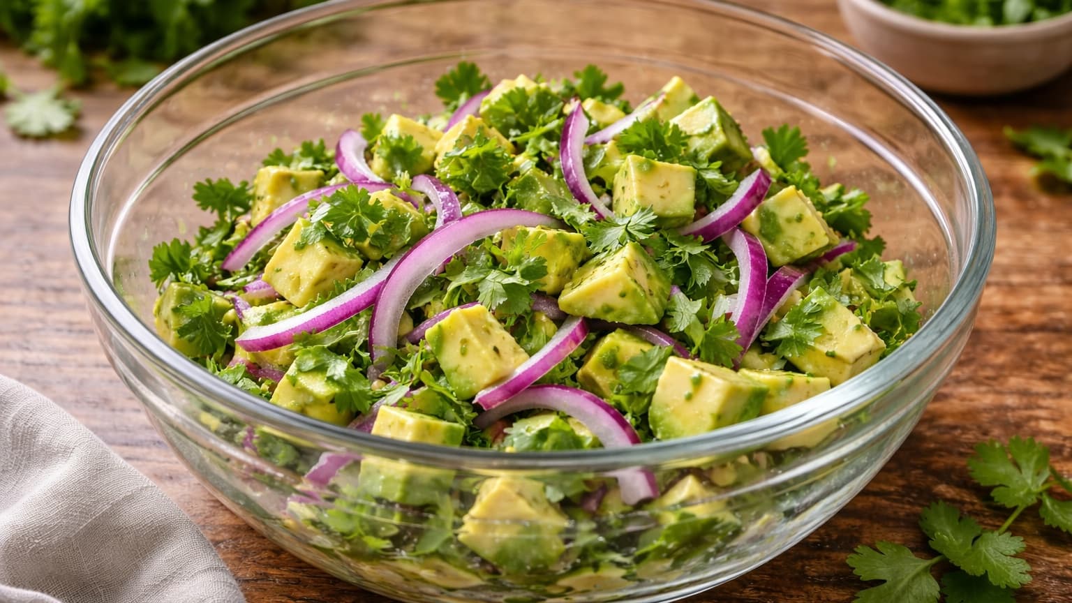 A mixing bowl filled with creamy green avocado cubes, thin purple-red onion slices, and fresh green cilantro. The ingredients are vibrant and fresh. The bowl sits on a wooden surface with soft daylight illuminating the colors.