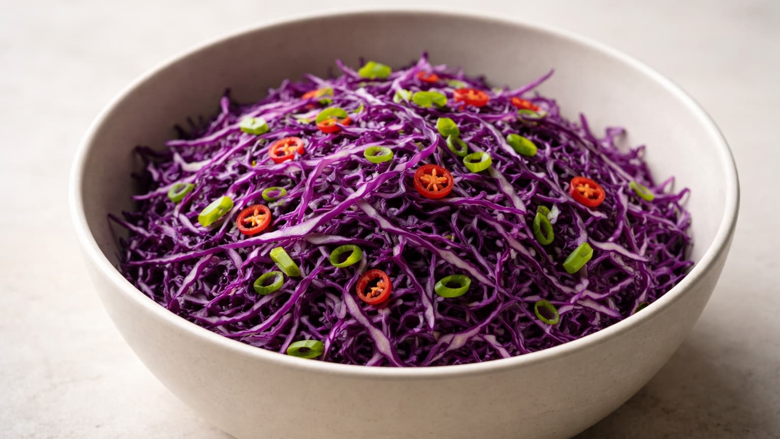 Minimalist food photograph shot from a 45-degree angle. A large matte ceramic bowl sits on a light neutral stone surface. Inside, finely shredded blue cabbage strands are piled loosely, thin and crisp, showing deep purple and blue hues. Thin red chili slices and bright green scallions are evenly distributed throughout. Soft diffused daylight from the left, neutral tones, shallow depth of field, no props, no background elements.