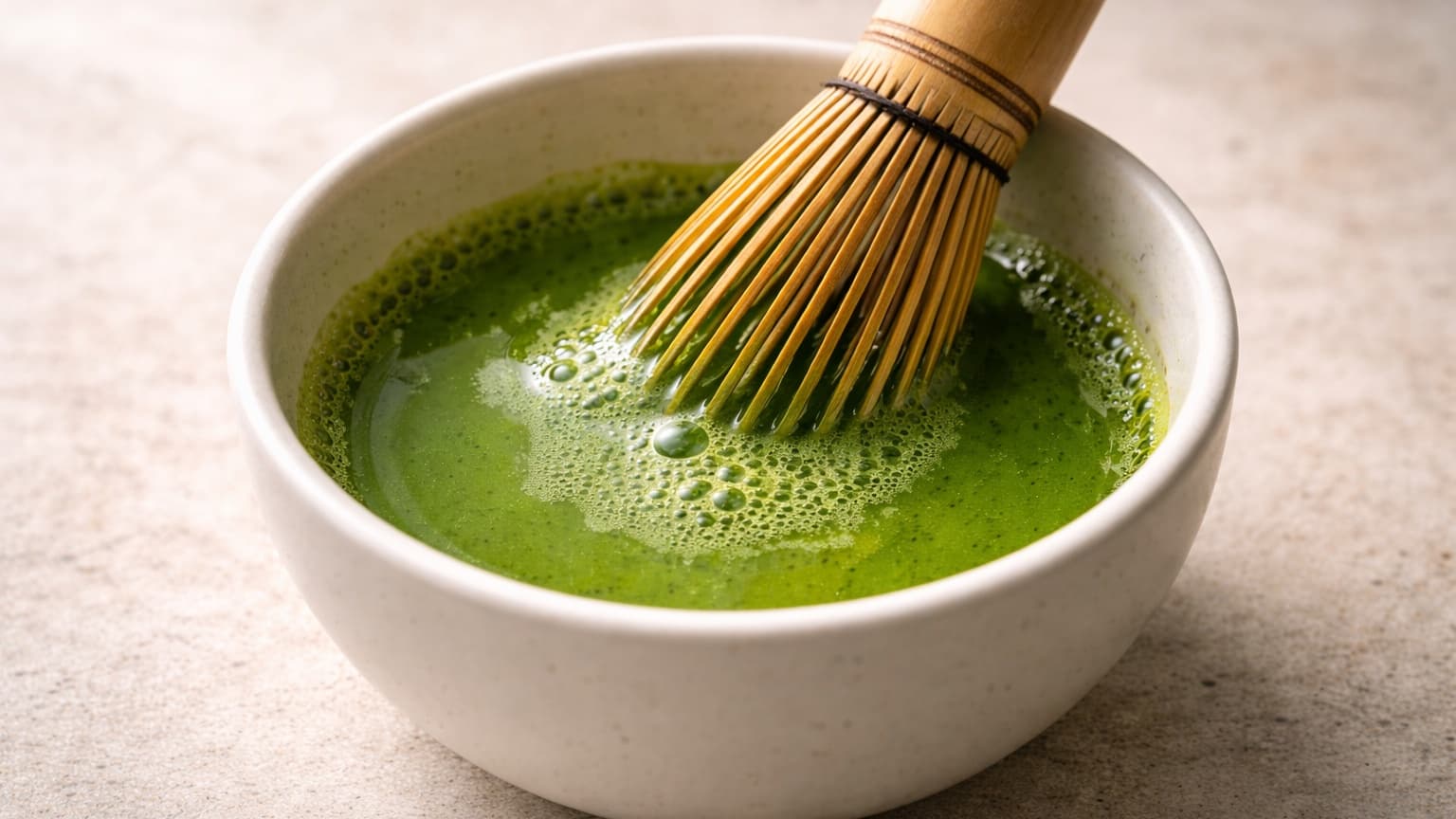 Close-up food photograph shot from a 45-degree angle. A small matte white ceramic bowl sits on a neutral stone surface. Inside, vivid green matcha tea is being whisked with a bamboo whisk, creating fine foam on the surface. The liquid is smooth and evenly colored with no clumps. Soft natural daylight from the left, shallow depth of field, minimal background with no props.