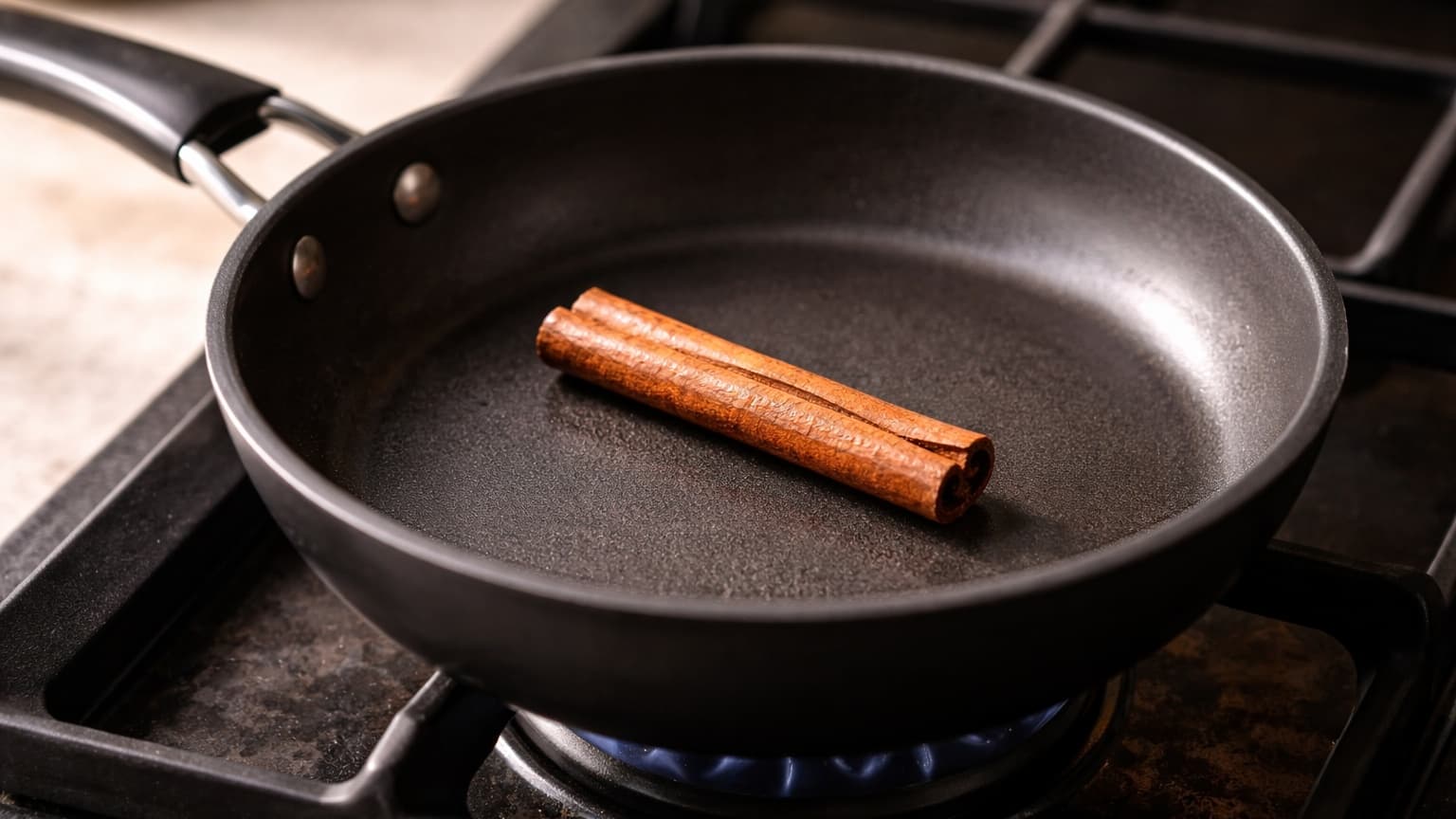 Minimalist kitchen photograph shot from a 45-degree angle. A small black pan sits on a stovetop with a single cinnamon stick gently warming inside. The cinnamon stick is dry, medium brown, and slightly glossy from heat. The background is softly blurred, warm natural light, realistic and subtle.