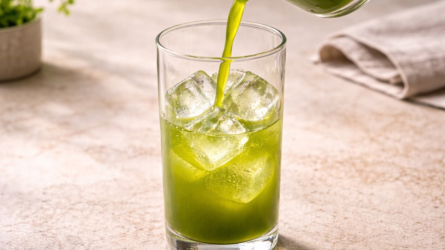 Natural food photograph shot from a 45-degree angle. A tall clear glass filled halfway with large, transparent ice cubes sits on a stone surface. A green liquid is being poured in, coating the ice and creating reflections. Clean, fresh look with neutral tones and soft daylight.