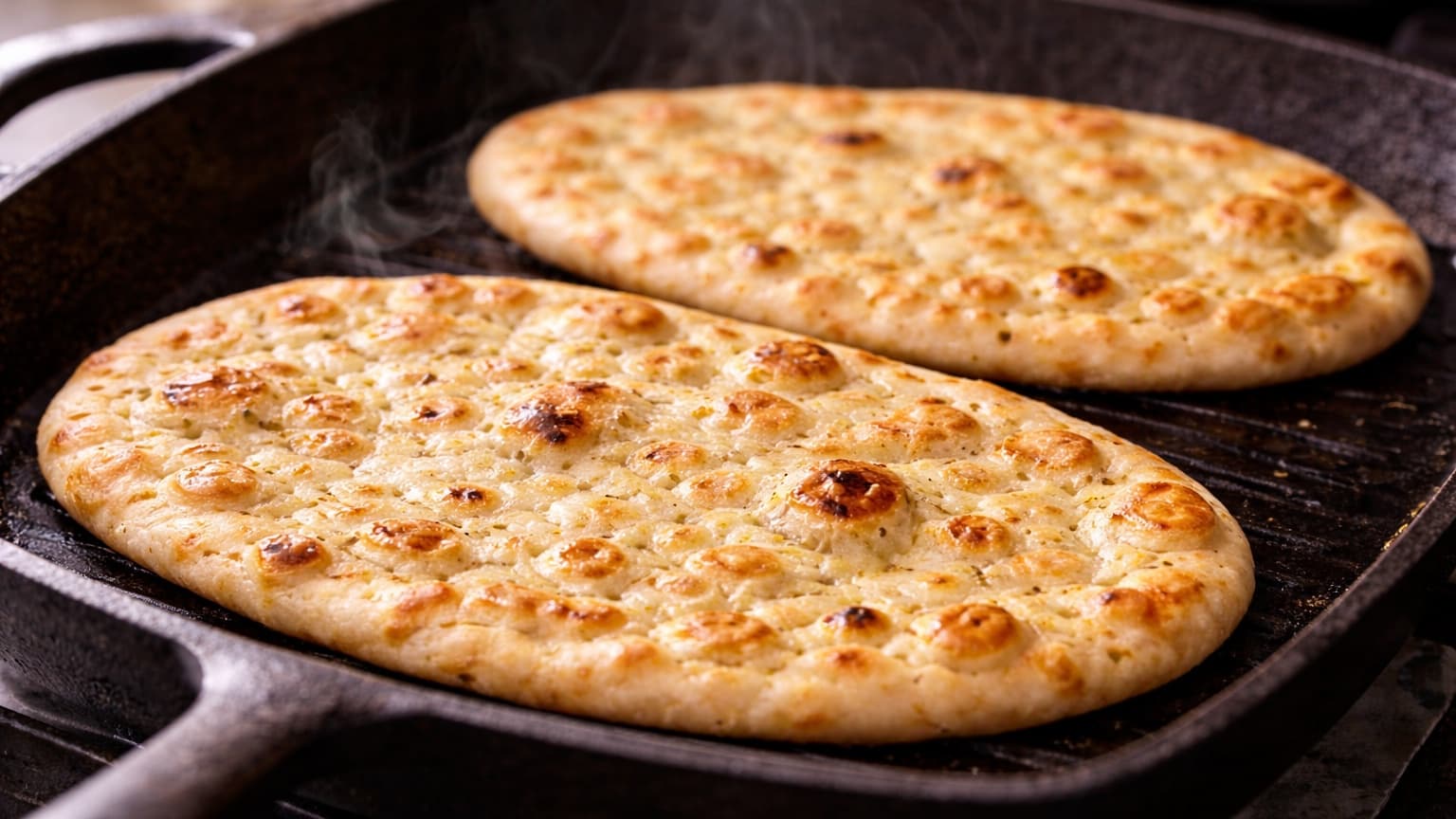 Close-up photograph at a slight angle of oval flatbreads warming in a cast iron pan. The bread surface has golden bubbles and light char spots. Gentle steam rises. Background kitchen scene is minimal and softly lit.