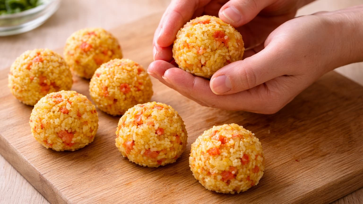 Close-up of hands shaping small round rice balls on a wooden cutting board. The rice balls are evenly sized, bright yellow with visible red kimchi bits. Soft natural kitchen lighting.