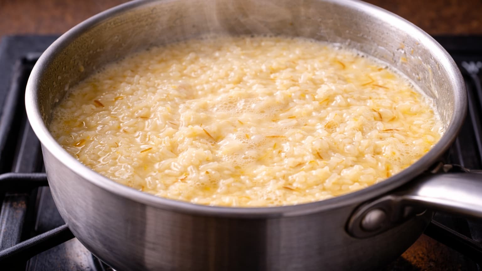 A medium stainless steel saucepan on a stovetop containing jasmine rice cooking in creamy coconut milk tinted light golden from saffron threads. The mixture is gently bubbling with steam rising. The rice grains are visible and partially submerged in the creamy liquid. Warm kitchen lighting highlights the pale yellow color and glossy surface of the cooking rice.