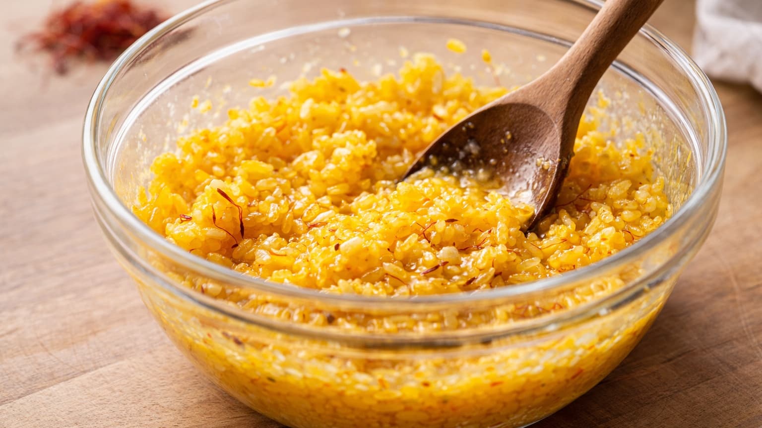 Close-up of a glass mixing bowl containing bright golden saffron rice being mixed with egg and cornstarch. The rice grains are glossy and yellow-orange from saffron. A wooden spoon stirs the mixture. The bowl sits on a light wooden kitchen countertop under soft natural daylight. Small saffron threads are visible throughout the rice mixture.