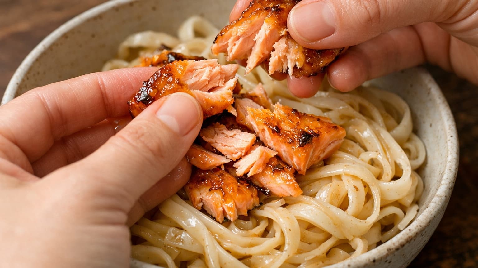 Hands gently flaking a glazed salmon fillet into tender pieces over a bowl of rice noodles. The salmon pieces are golden on the outside with pink flaky interiors. The noodles underneath look silky and lightly coated with sauce.